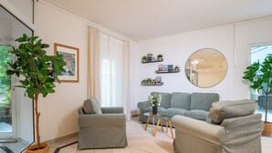 A modern living room with a grey sofa, a small armchair, and a coffee table. There are potted plants and a round mirror on the walls. - Waldhotel Schöneiche - Self-Check-in (Schoeneiche bei Berlin)