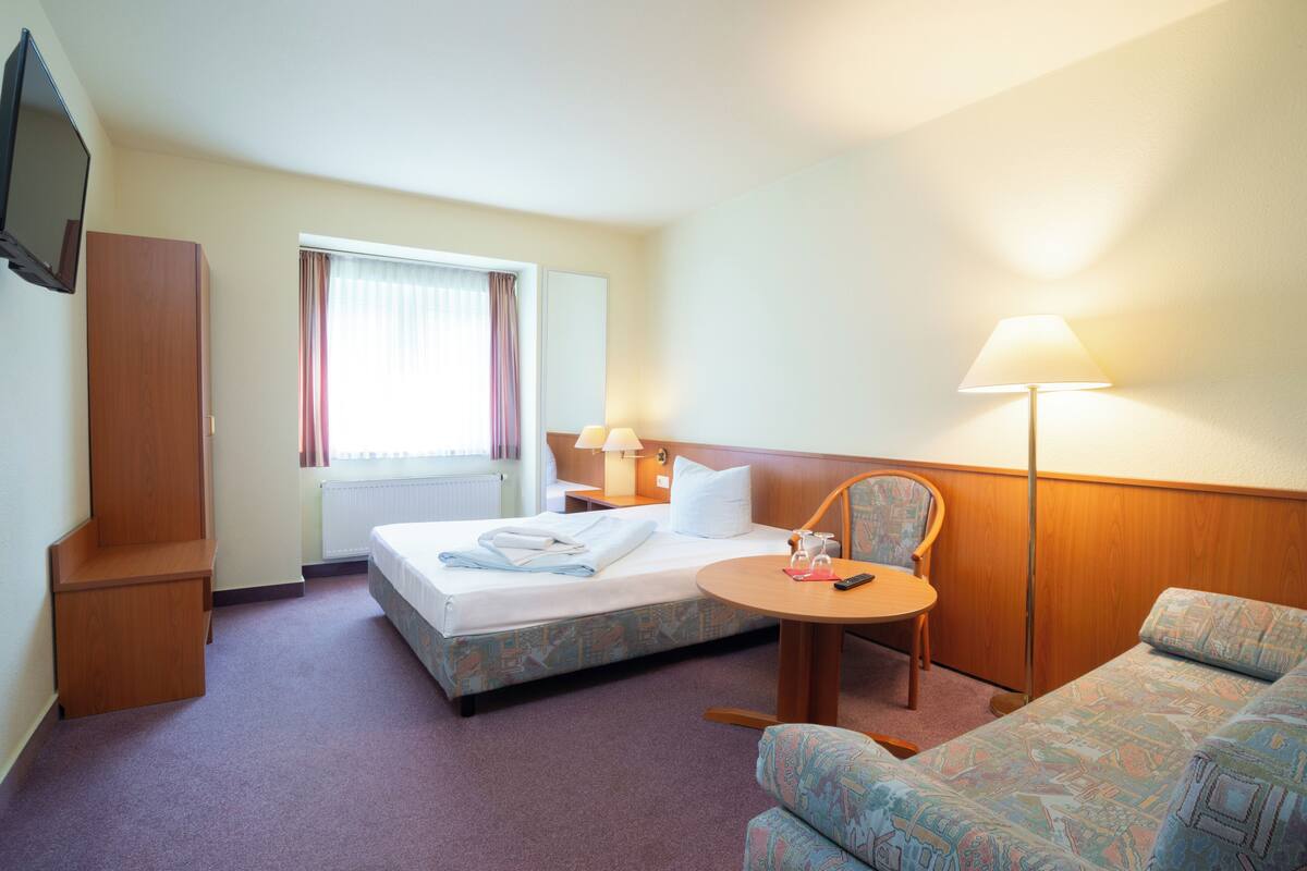 A hotel room with a bed, a desk with a lamp, a chair, and a sofa.
