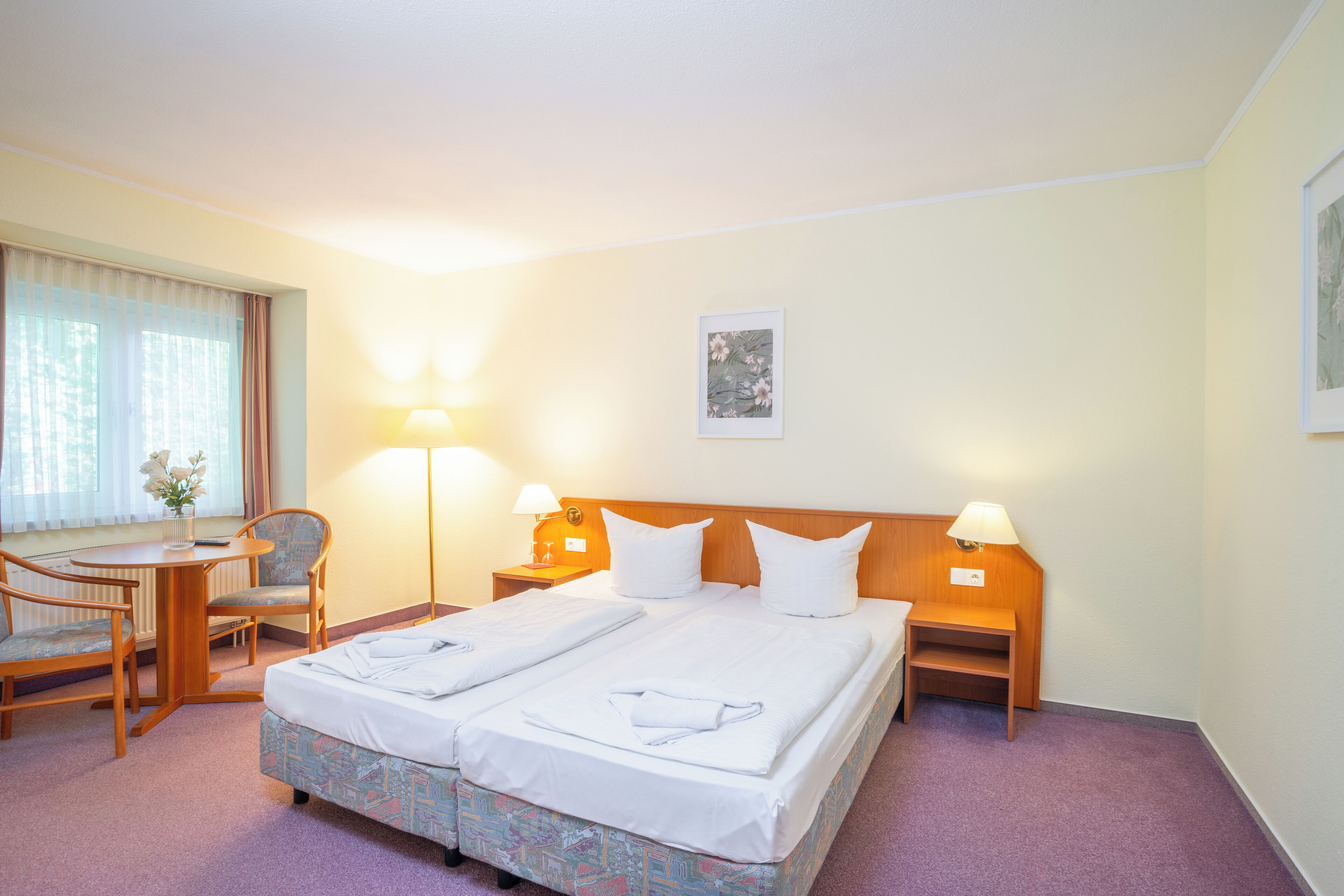 Quarto de hotel com cama de casal, duas mesas de cabeceira com luminárias, uma pequena escrivaninha com cadeira e uma janela com cortinas.