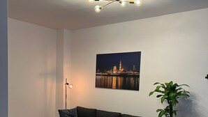 A modern living room with a dark sofa, a coffee table, a floor lamp, a potted plant, and a cityscape painting on the wall.