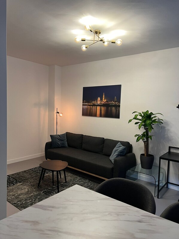 A modern living room with a dark sofa, a coffee table, a floor lamp, a potted plant, and a cityscape painting on the wall.