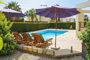 A swimming pool with lounge chairs and umbrellas, surrounded by greenery and a stone wall. - Villa Coralia Dream 12 (Pegeia)