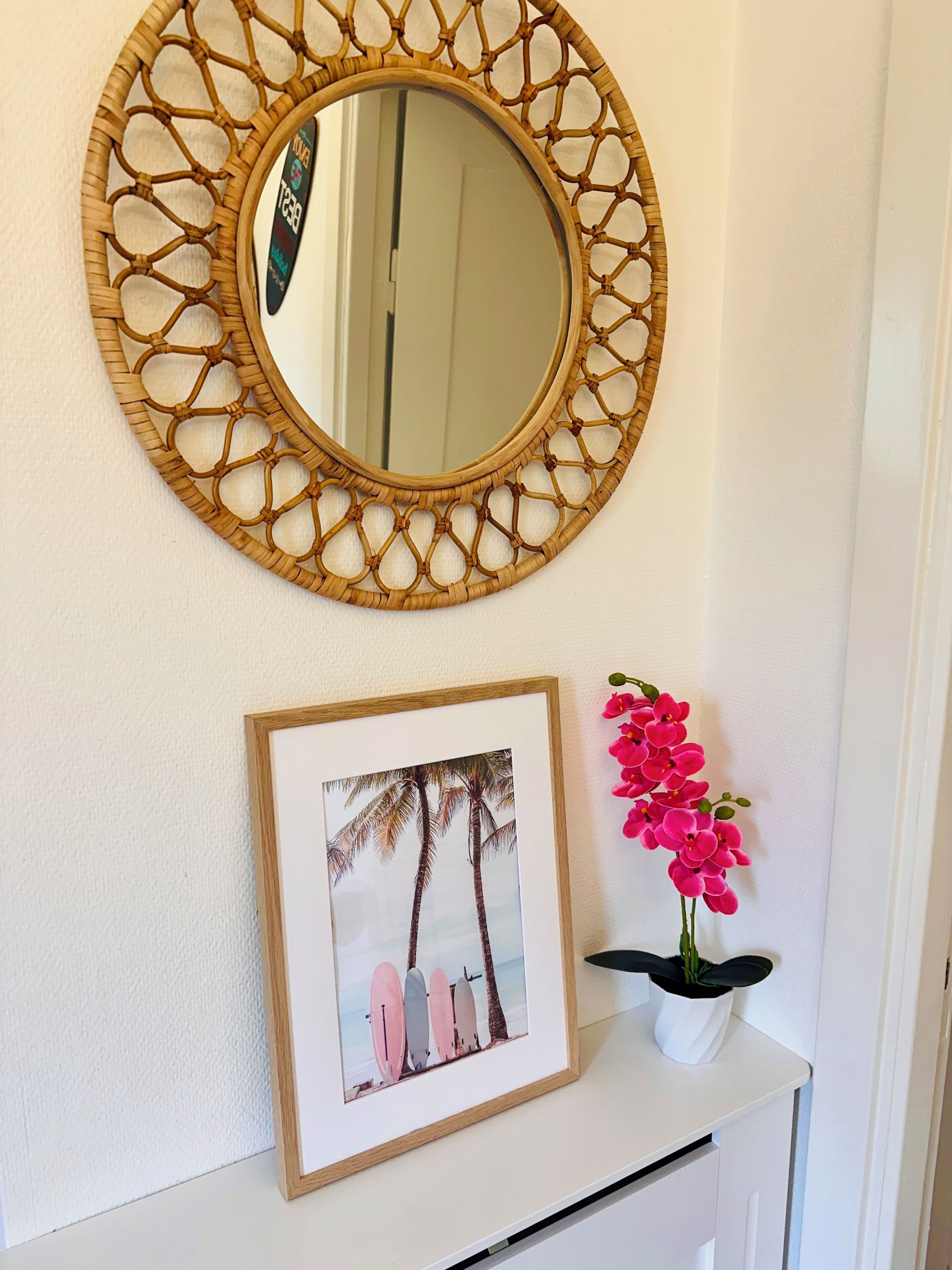 A wall with a woven mirror, a framed picture of surfboards, and a potted orchid.
