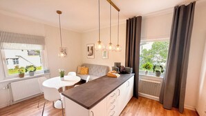Una cocina moderna con una isla central, luces colgantes, una zona de comedor con mesa y sillas blancas, y vistas al exterior a través de amplios ventanales.