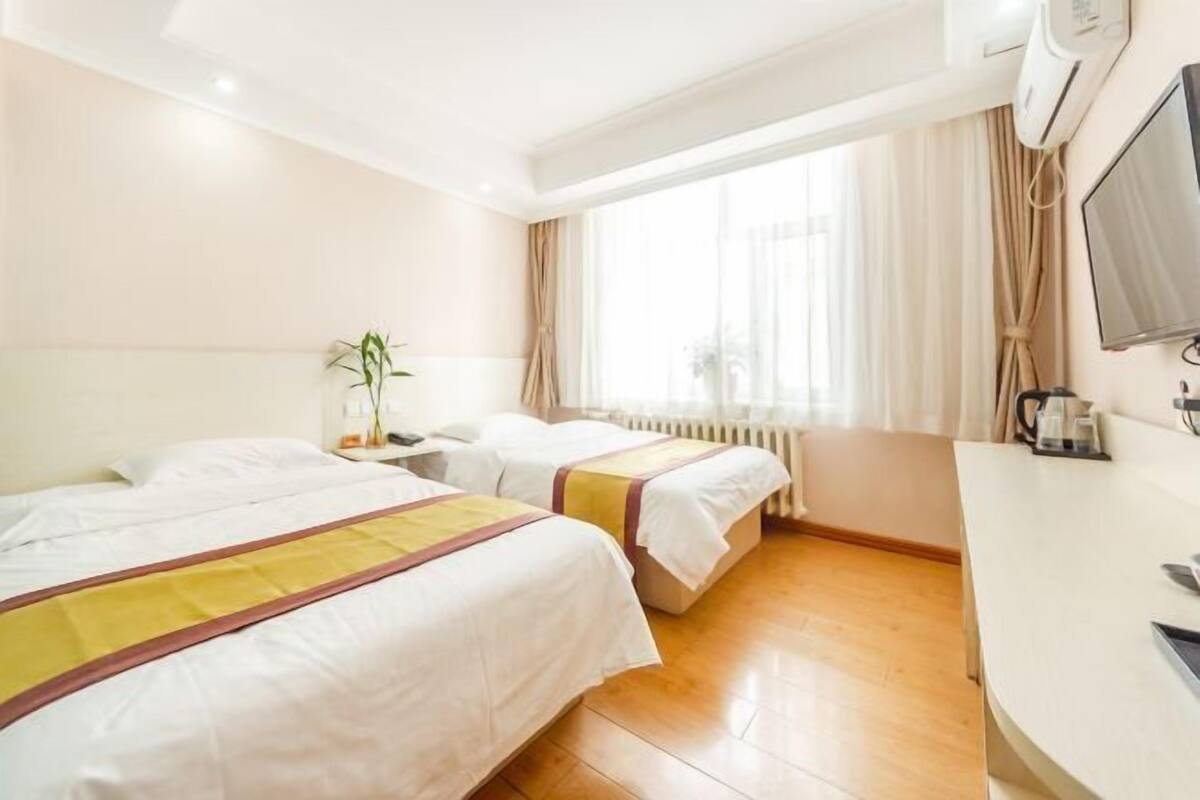 A hotel room with two beds, a desk, a radiator, and a window with curtains.