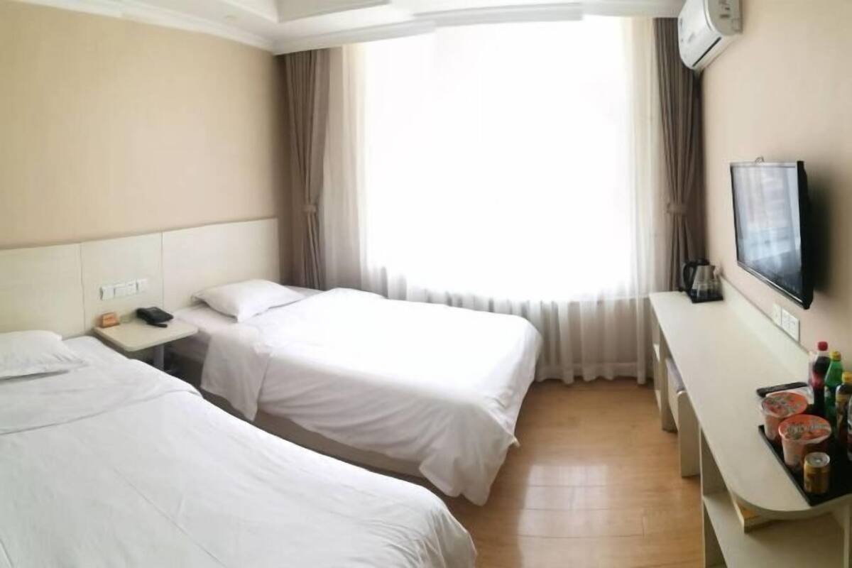 A hotel room with two single beds, a desk with a flat-screen TV, and a window with curtains.