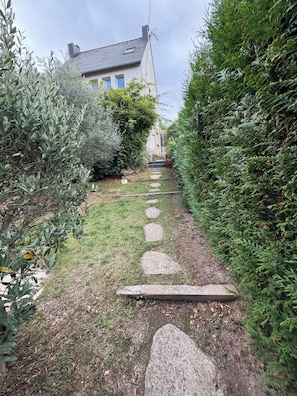 Ein steiniger Weg führt zu einem Haus mit geneigtem Dach, umgeben von üppiger Vegetation.
