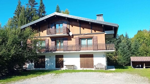 A two-story wooden house with a balcony, surrounded by trees.