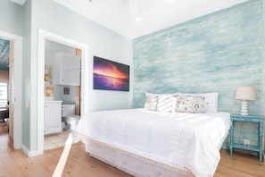 A bedroom with a bed, a painting, a nightstand, and a bathroom visible through an open door. - Sandy Coral (Port Aransas)