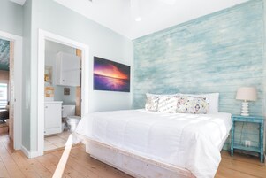 Ein Schlafzimmer mit Bett, einem Bild, einem Nachttisch und einem durchgegangenen Türspalte erkennbaren Badezimmer.
