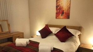 A neatly made bed with a plaid blanket and two red pillows, a bedside table with a lamp, and a painting of a couple on a bridge. - ExploreNESS Apartment  (Inverness)