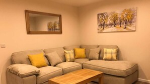 A living room with a beige sectional sofa, a wooden coffee table, a mirror, and a painting of trees. - ExploreNESS Apartment  (Inverness)