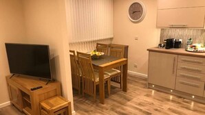 A compact living space with a TV, wooden dining table, chairs, a small wooden stool, a kitchen area with cabinets, and a clock on the wall. - ExploreNESS Apartment  (Inverness)