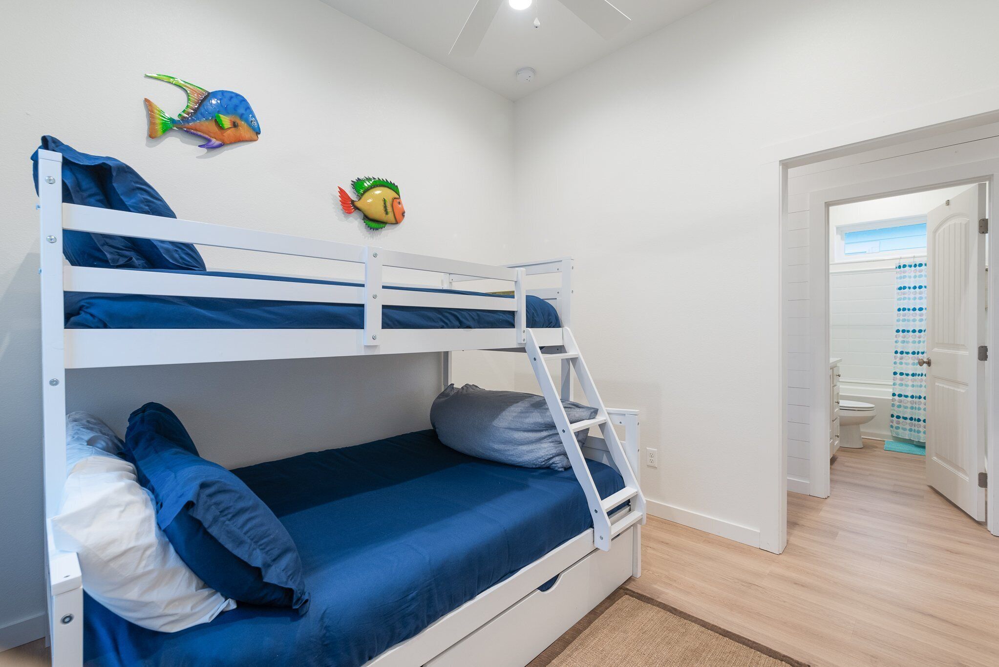 A bunk bed room with a ladder, a blue mattress, and pillows. There is a fish decoration on the wall and a bathroom visible through an open door.