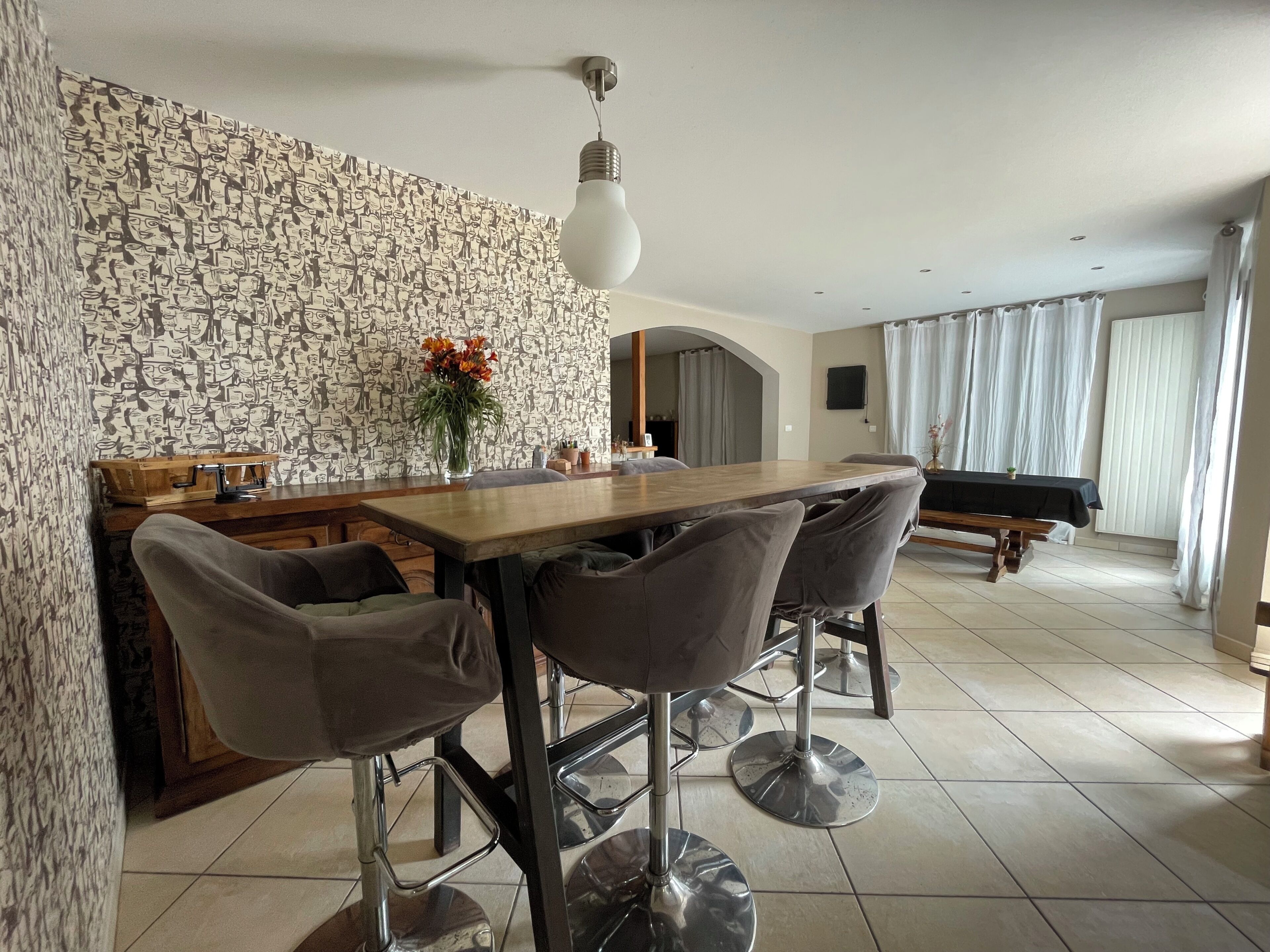 A dining area with a wooden table and four bar stools, a stone-patterned wall, a vase with flowers, and a ceiling light fixture.