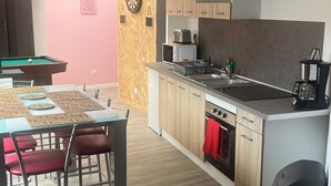 Une cuisine moderne avec des meubles en bois, un four encastré et une machine à café. On y trouve également un coin repas avec un billard et des chaises rouges.