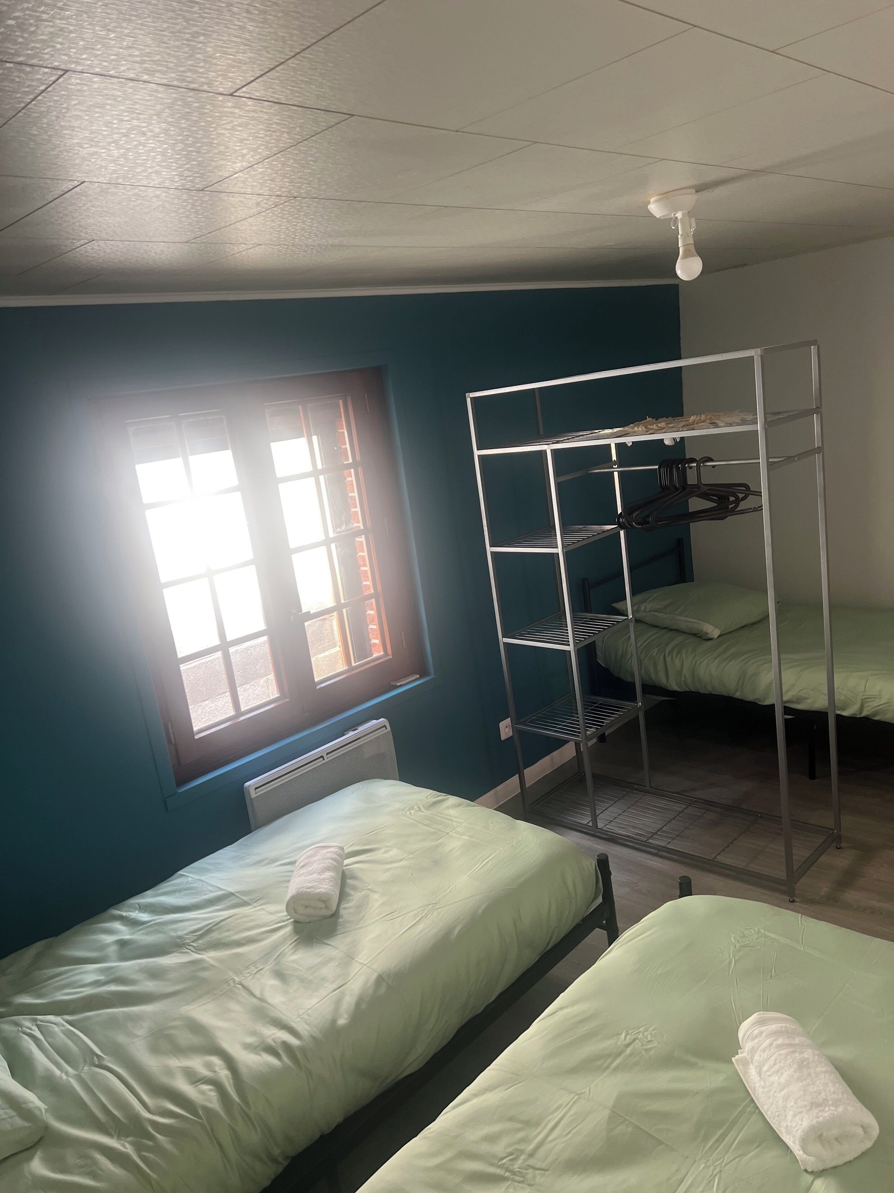 Een studentenkamer met twee eenpersoonsbedden, een stapelbed, een radiator en een raam met zonwering.