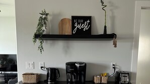 Keittiössä on musta työtaso, valkoiset kaapit, Keurig-kahvinkeitin, leivänpaahdin ja kyltti, jossa lukee: ”OLE HYVÄ JA VIIVY”.