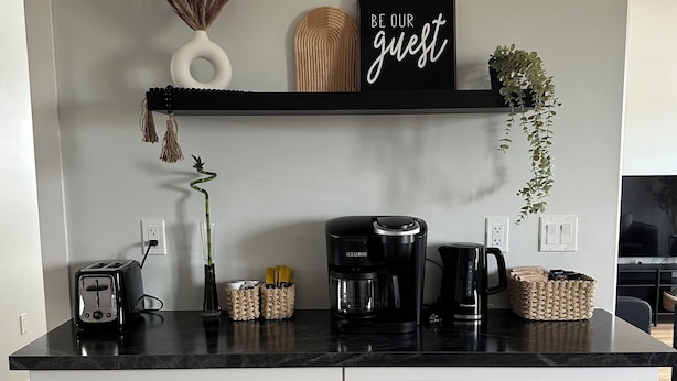 A kitchen with white cabinets, a black countertop, a Keurig coffee maker, a toaster, and a sign that says "BE OUR guest".