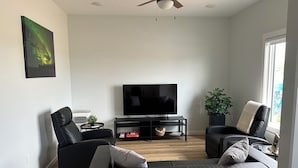 A modern living room with a flat-screen TV, two black sofas, a ceiling fan, and a large window.