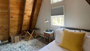 A cozy attic bedroom with wooden beams, a bed with white and yellow bedding, a small table with a lamp, and a window with a view. - Honest Abe Frame @ Sunrise (Ellicottville)