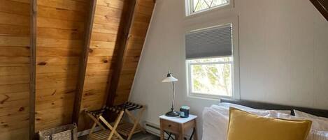 A cozy attic bedroom with wooden beams, a bed with white and yellow bedding, a small table with a lamp, and a window with a view.