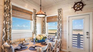 A dining area with a wooden table set for four, a hanging pendant light, and a view of the beach through the windows. - Jetty Set Go! Ocean Views & Private Pool! (Port Aransas)