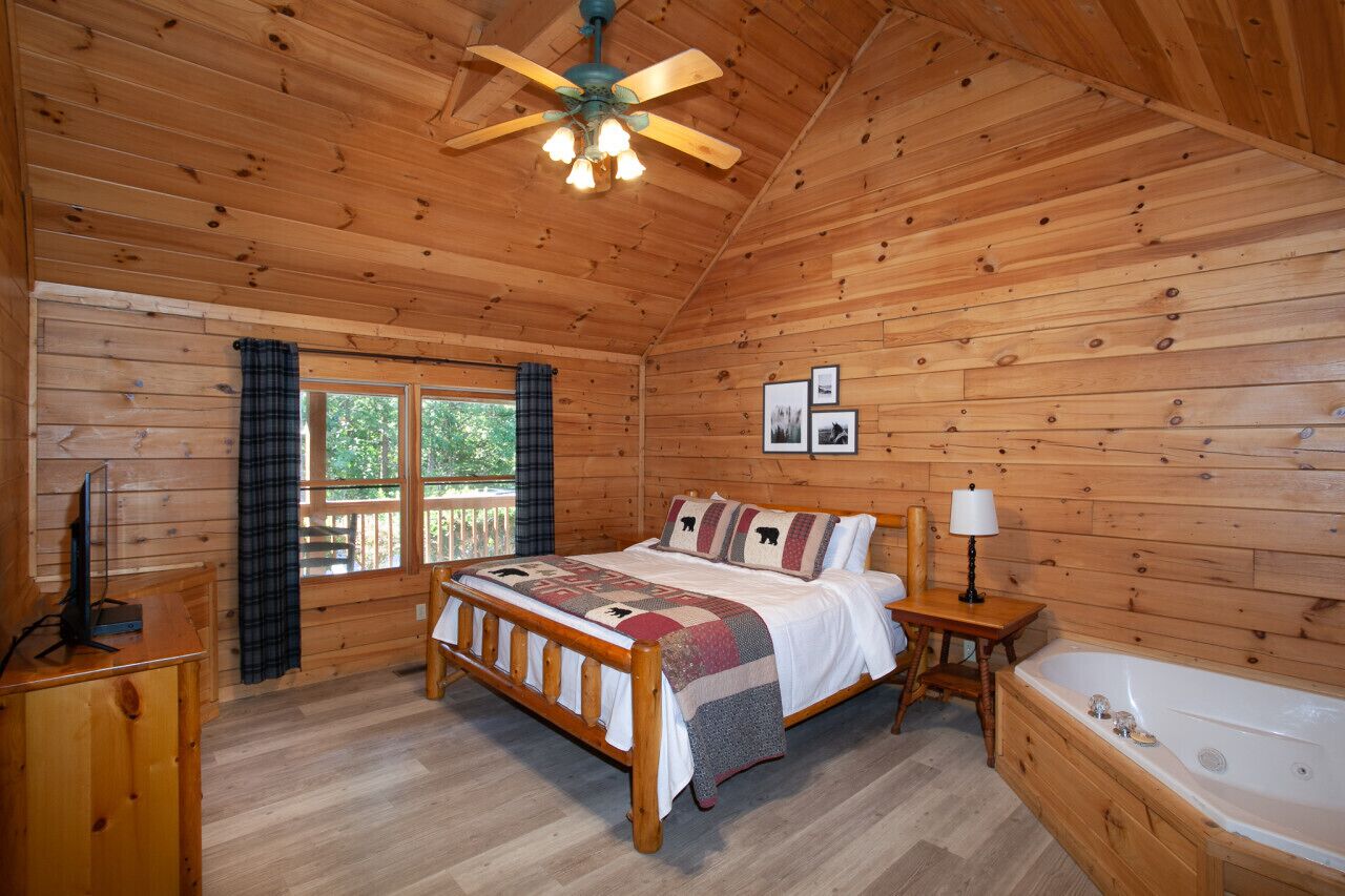 A wooden cabin bedroom with a bed, a nightstand, a lamp, a window with curtains, and a ceiling fan.