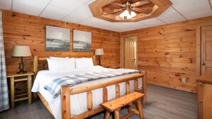 Un dormitorio con paneles de madera, una cama, dos mesitas de noche, un banco y un ventilador de techo.