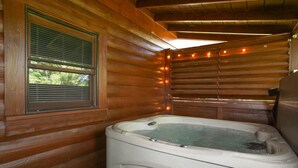 Interior de una cabaña de madera con un jacuzzi, una ventana con persianas e iluminación de luces colgantes.