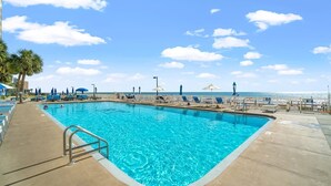 A rectangular swimming pool with clear blue water, surrounded by lounge chairs and umbrellas, with a view of the ocean and palm trees in the background. - Prancing Peacock Gulf View Condo (Panama City Beach)