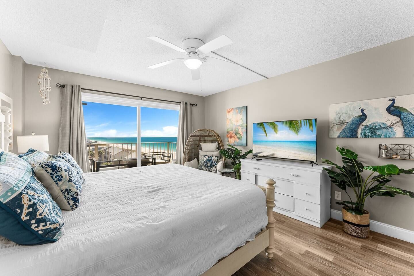 A bedroom with a large bed, a ceiling fan, a TV, and a view of the beach.