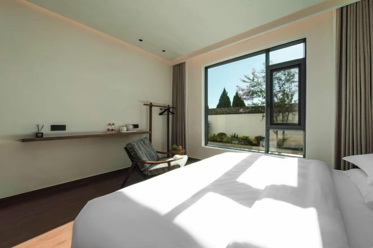 A modern hotel room with a large bed, a chair, a desk, and a window with a view of trees.