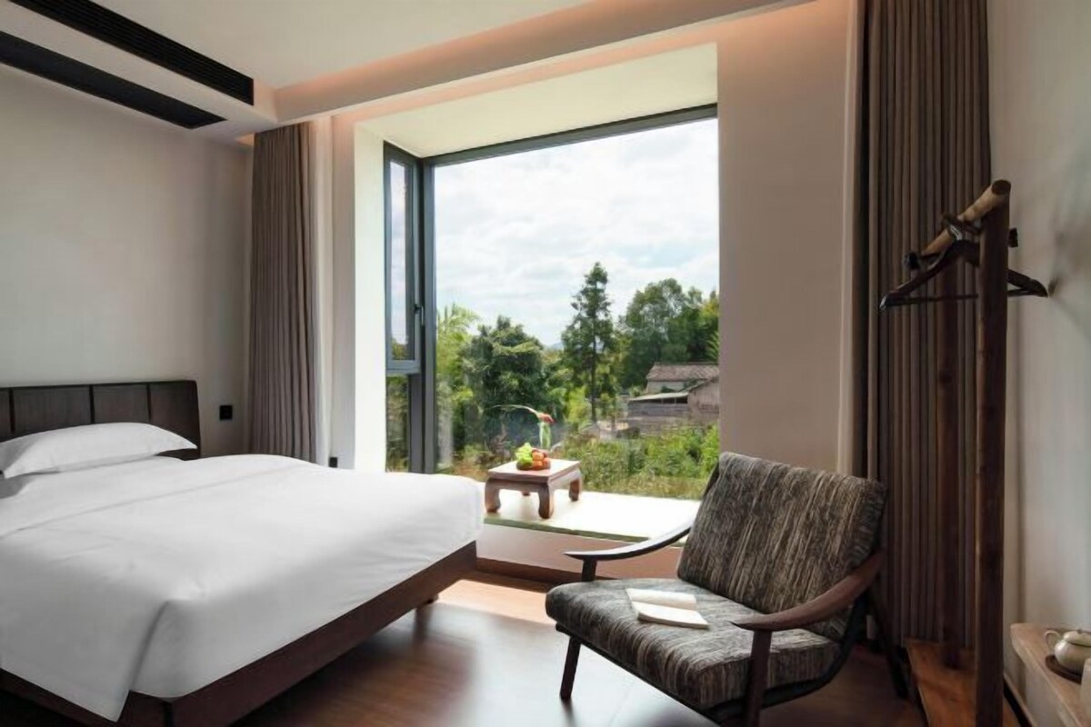 Ein Hotelzimmer mit einem Bett, einem Sessel, einem kleinen Tisch und Blick auf Grünflächen.