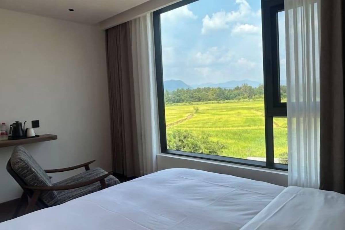 A hotel room with a large window, a bed, a chair, and a shelf with items.