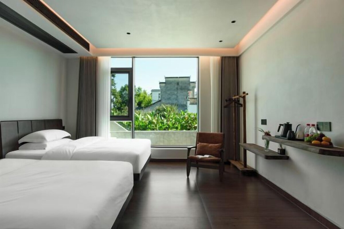Ein Hotelzimmer mit zwei Betten, einem Sessel, einem kleinen Tisch und Blick auf Grünflächen.