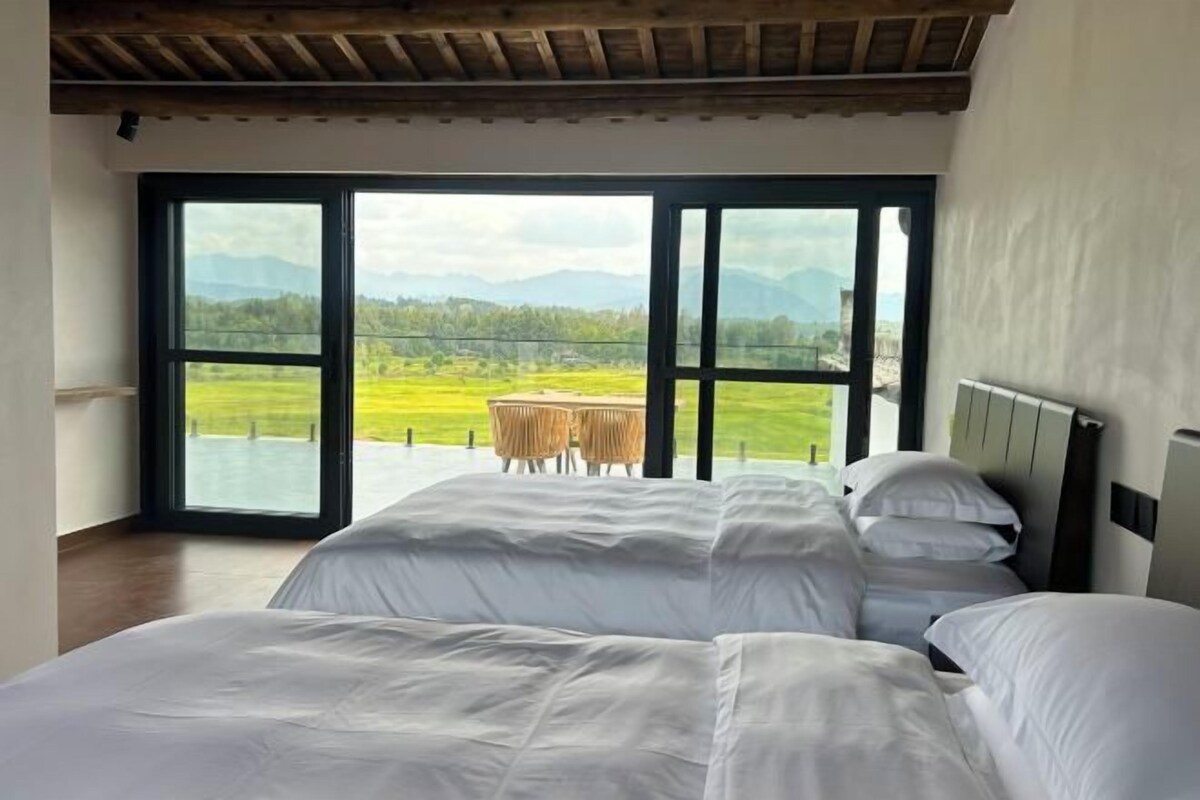 A bedroom with a large bed, a view of a green landscape through a sliding door, and a table with chairs outside.