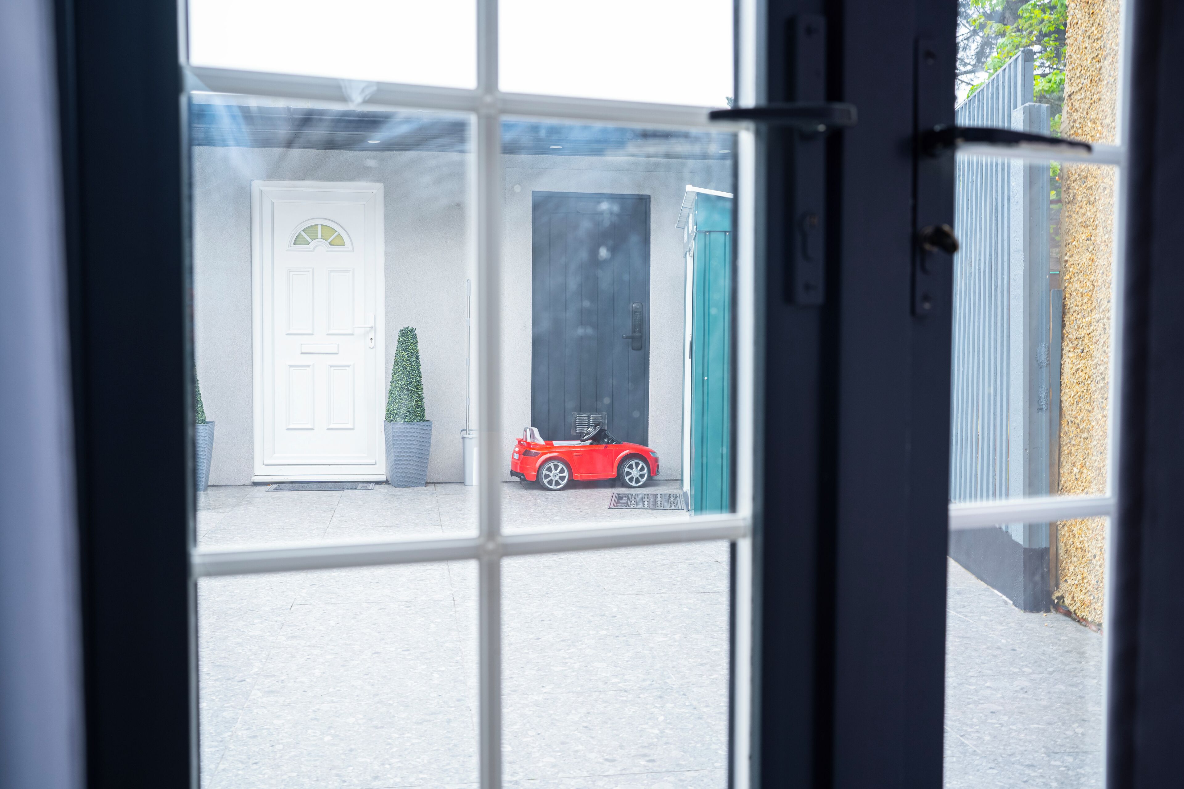 A view through a window of a building with a white door, a dark door, and a red car parked outside.