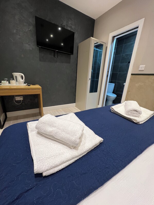 A hotel room with a bed, a desk with a kettle, cups, and a remote control. There is a TV mounted on the wall and a bathroom visible through an open door. - Rest Inn Edmonton (London)