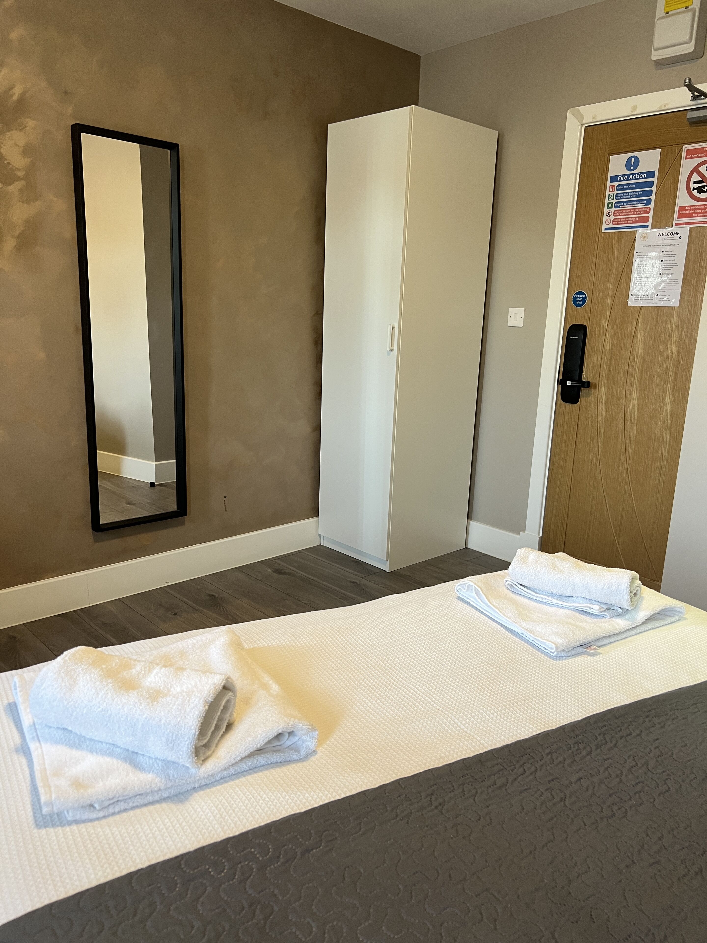 A hotel room with a bed, a mirror, a wardrobe, and a door with a fire action sign.