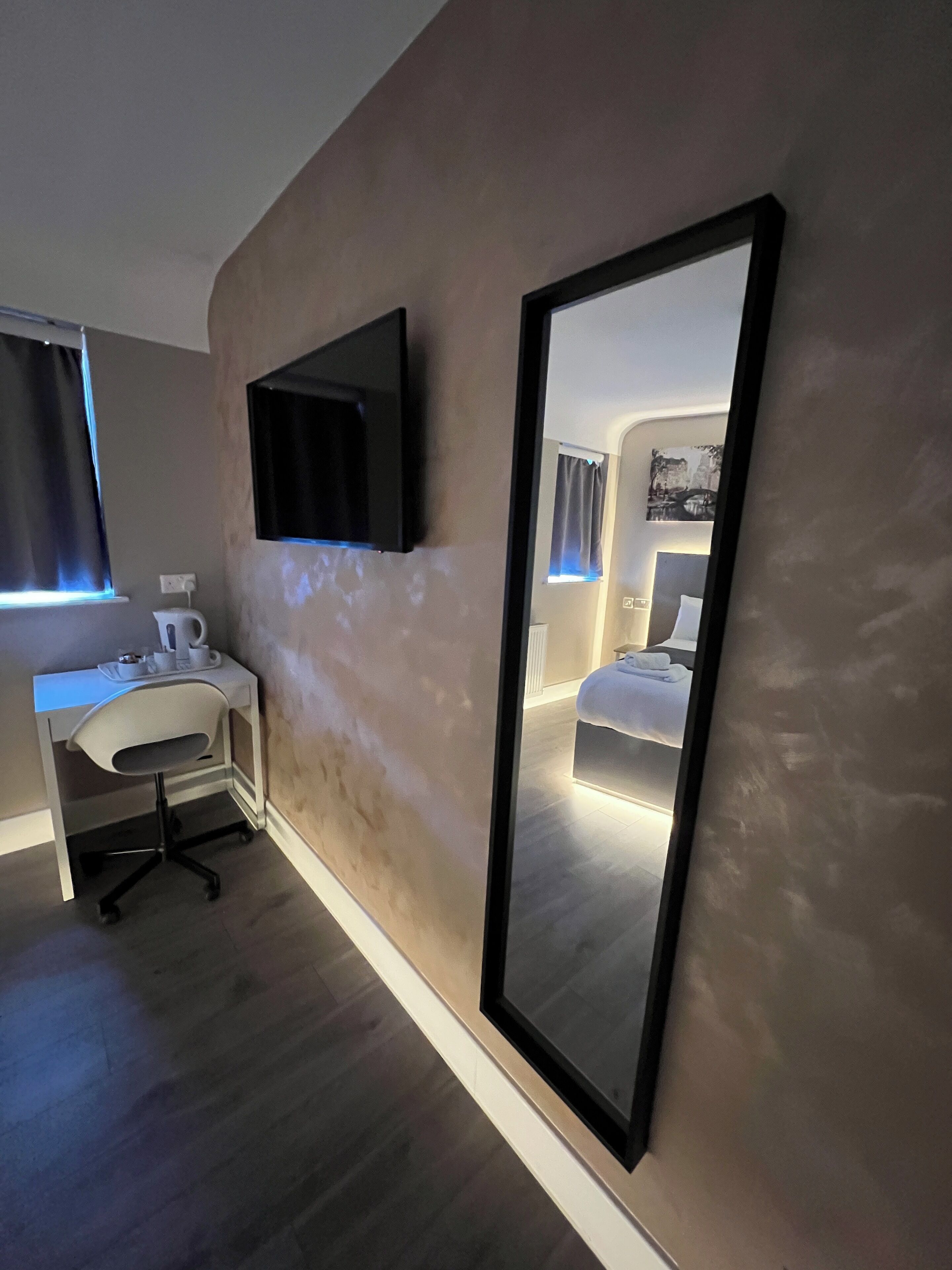 Una habitación de hotel moderna con un espejo grande, un televisor de pantalla plana, un escritorio con silla y una cama con cabecera.