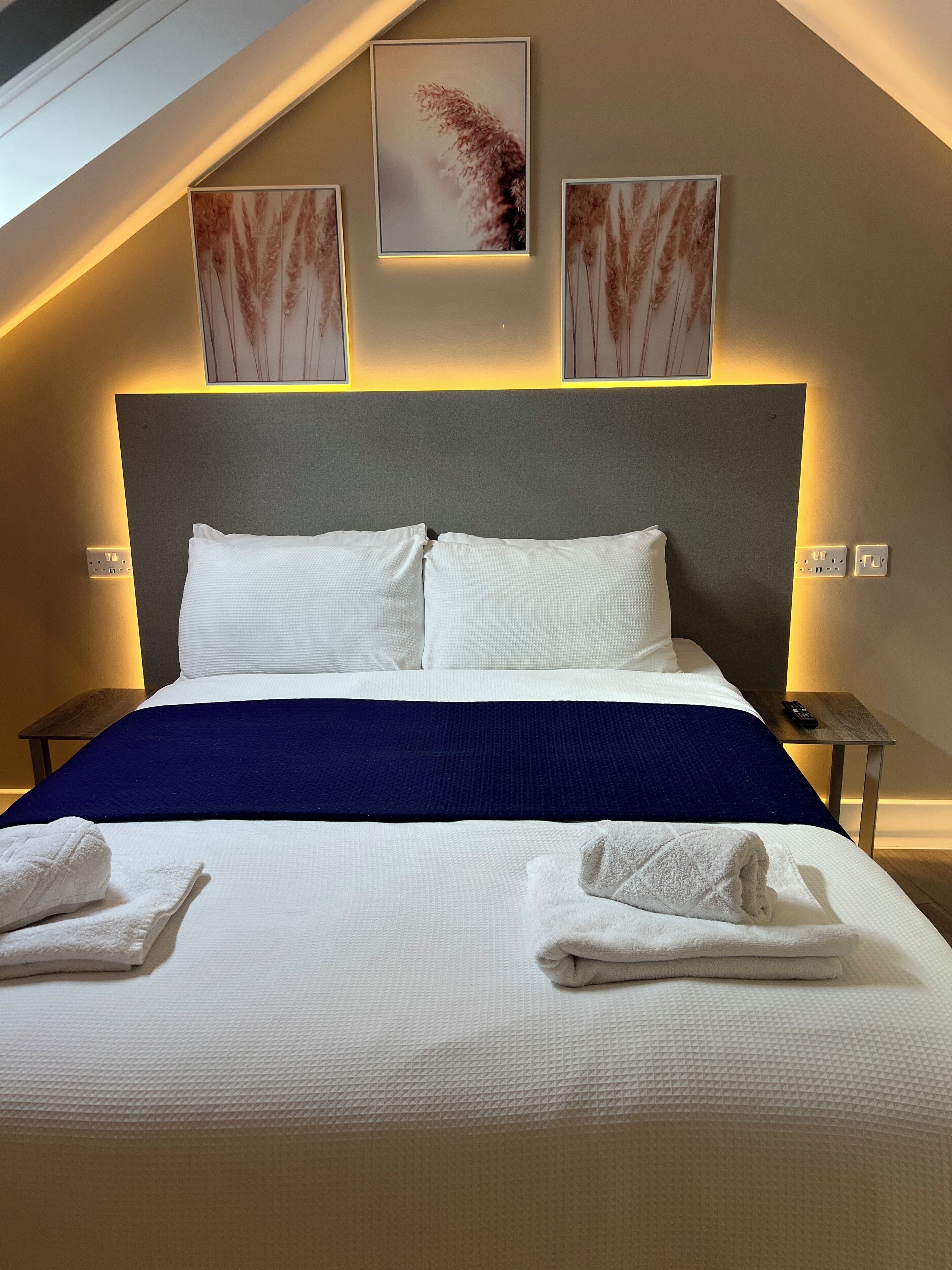 A neatly made bed with white linens and a blue accent, two white towels on the bed, and a remote control on a wooden side table.