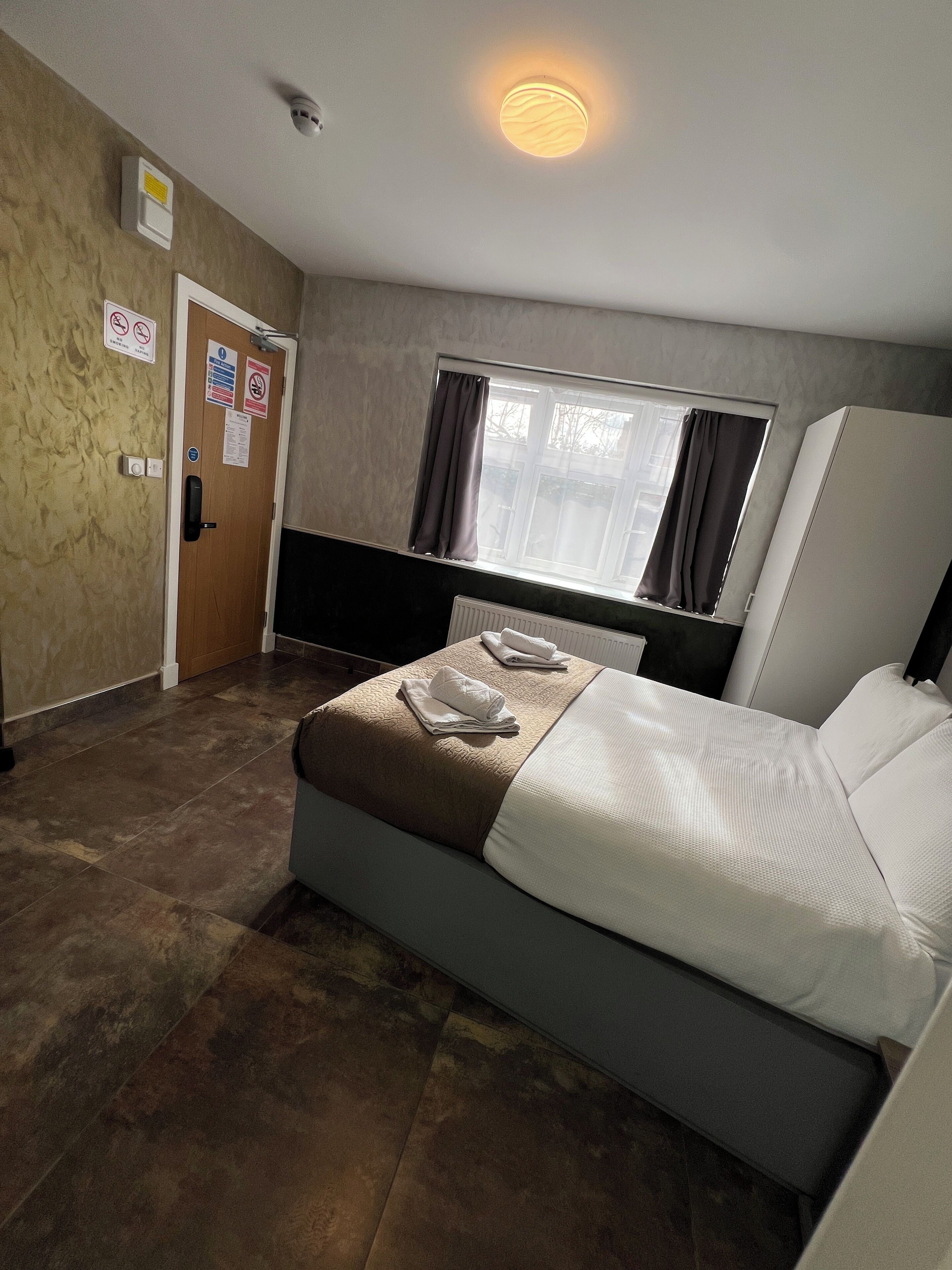 Une chambre d’hôtel avec un lit, une fenêtre avec des rideaux, un radiateur, une porte avec une pancarte et un luminaire au plafond.
