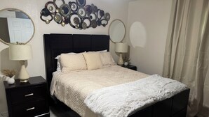 Un dormitorio con una cama negra, ropa de cama blanca, dos mesitas de noche con lámparas, un adorno en la pared y una ventana con cortinas.