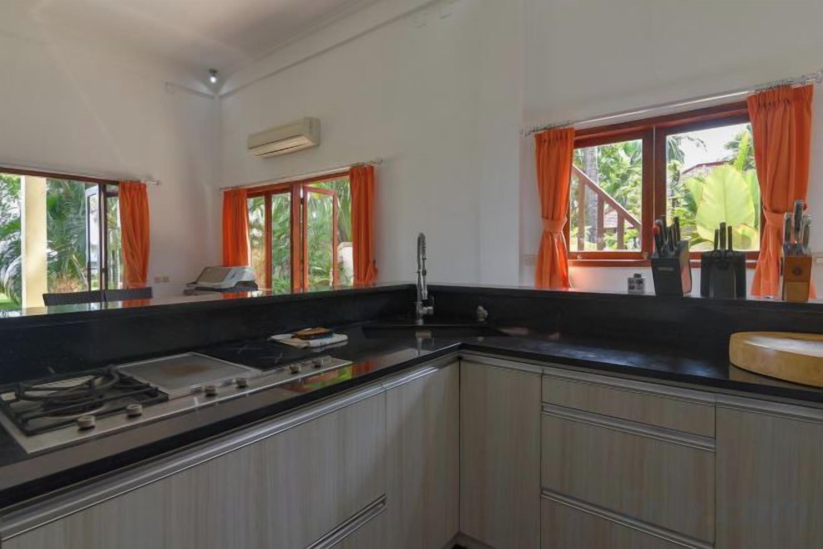A kitchen with a gas stove, sink, and cabinets. There are windows with orange curtains and a view of greenery outside.