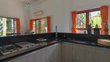 A kitchen with a gas stove, sink, and cabinets. There are windows with orange curtains and a view of greenery outside.