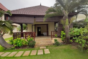 A two-story house with a tiled roof, a front porch, and a garden with palm trees and shrubs. - Villa Singkenken (Bali)