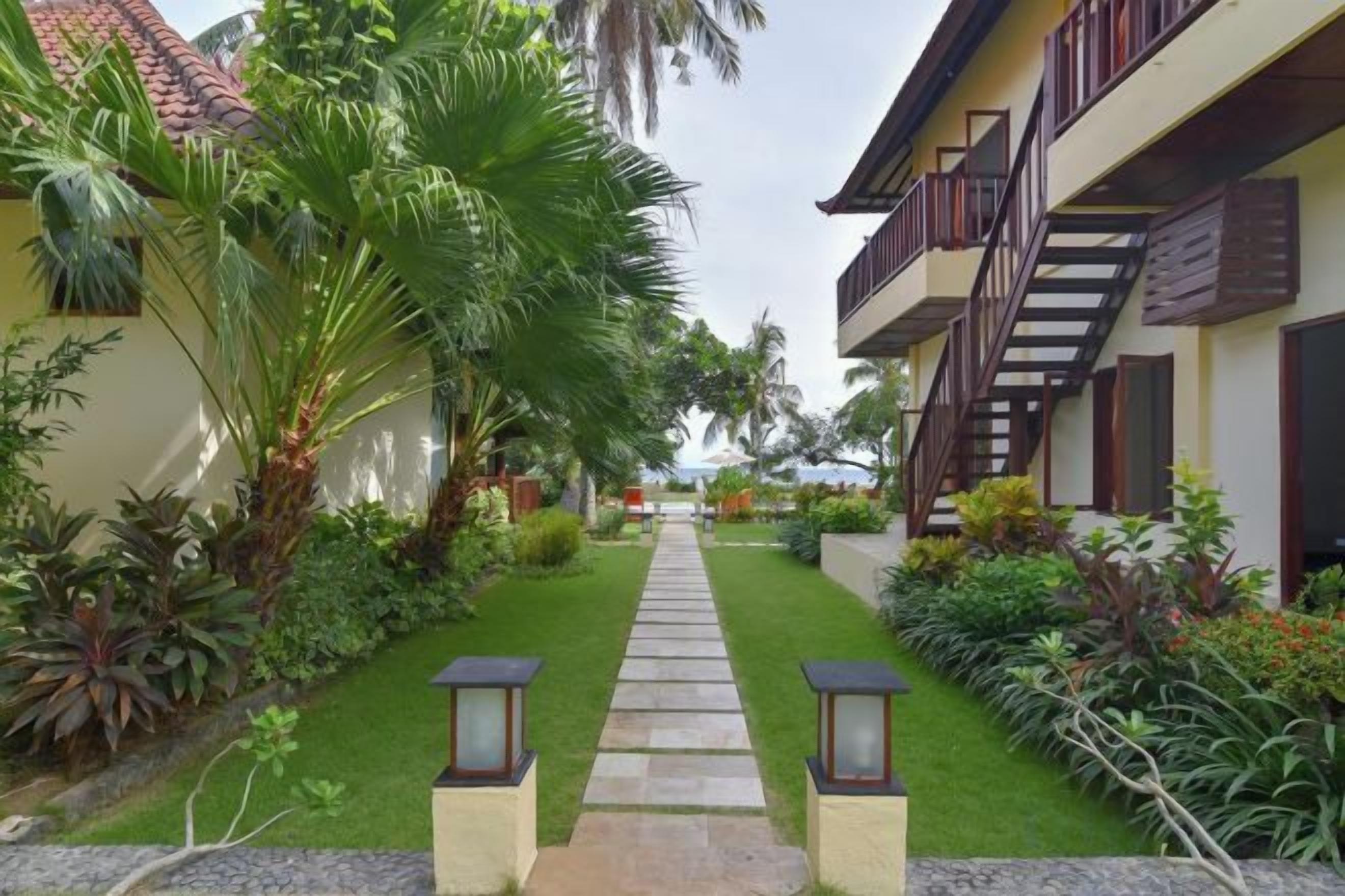 A pathway leading to a building with a staircase, surrounded by lush greenery and palm trees.