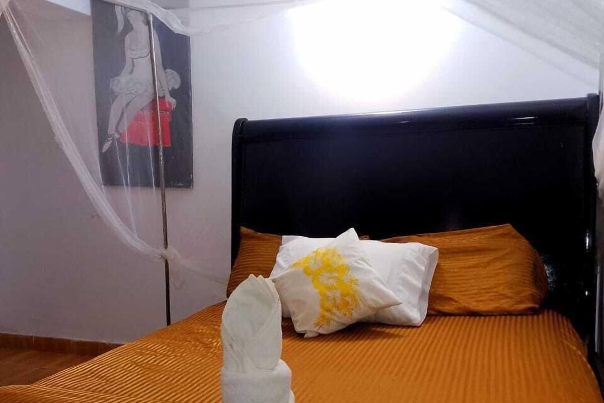 Una cama bien tendida con colcha naranja y almohadas blancas, cabecera negra y un cuadro en la pared.
