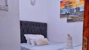 Une chambre à coucher avec une tête de lit rembourrée de couleur grise, un lit avec une literie blanche et une couverture à rayures, un tapis rouge et un tableau représentant des éléphants accroché au mur.
